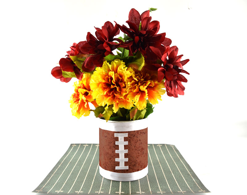 DIY football vase centerpiece made from a recycled tin can with white ribbon laces and vibrant red and yellow flowers.
