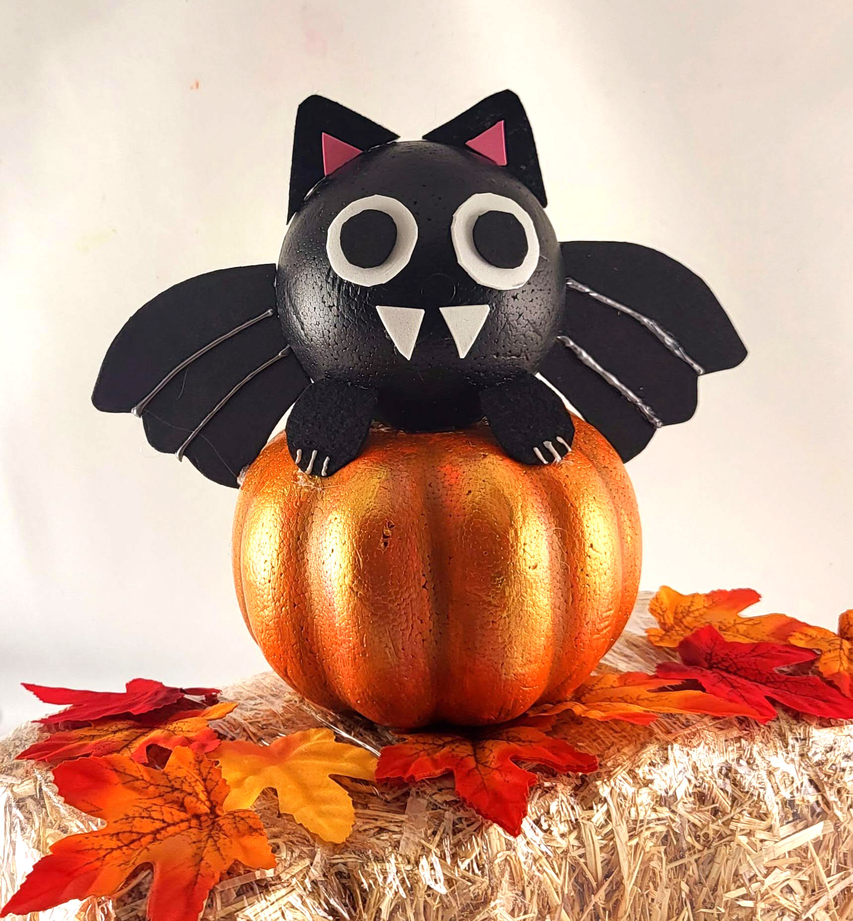 This Adorable DIY Dollar Tree foam pumpkin bat on a small hay bale. The cute black bat sits on a metallic orange pumpkin, surrounded by autumn leaves, creating a perfect Halloween decor scene.
