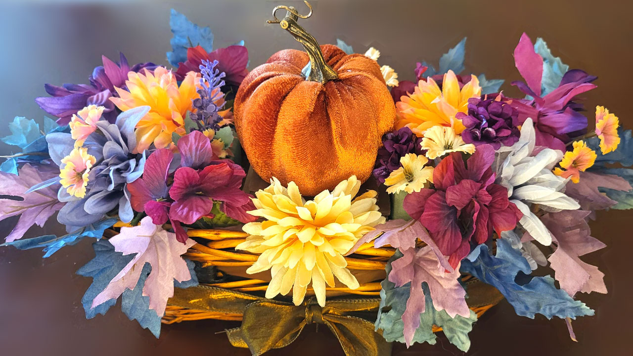 A soft orange velvet pumpkin sits as the centerpiece in a rustic wicker basket, surrounded by a vibrant fall floral arrangement of yellow, deep red, and purple flowers with unique blue autumn leaves.