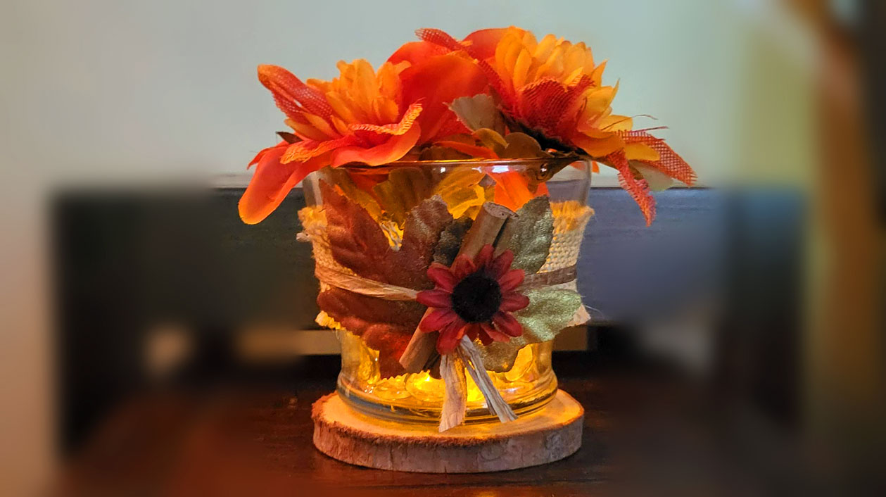 A high-end DIY fall candle holder made with Dollar Tree items, featuring orange mums, fall leaves, and a cinnamon stick on a rustic wood slice base.