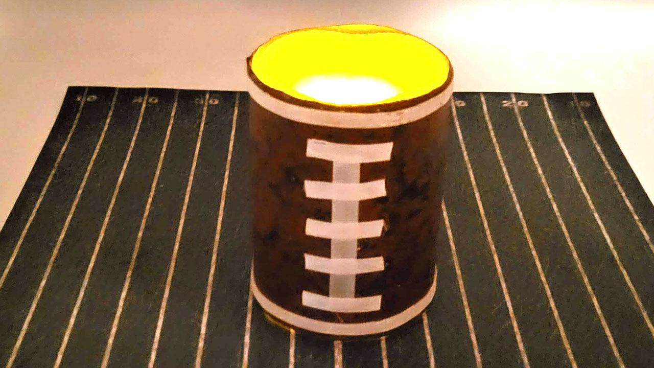 A close-up of a football candle glowing with a warm orange light on a green felt football field background.