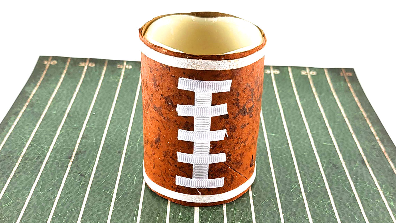Football candle made from a brown-painted cardboard roll with white ribbon laces, sitting on a football field mat.