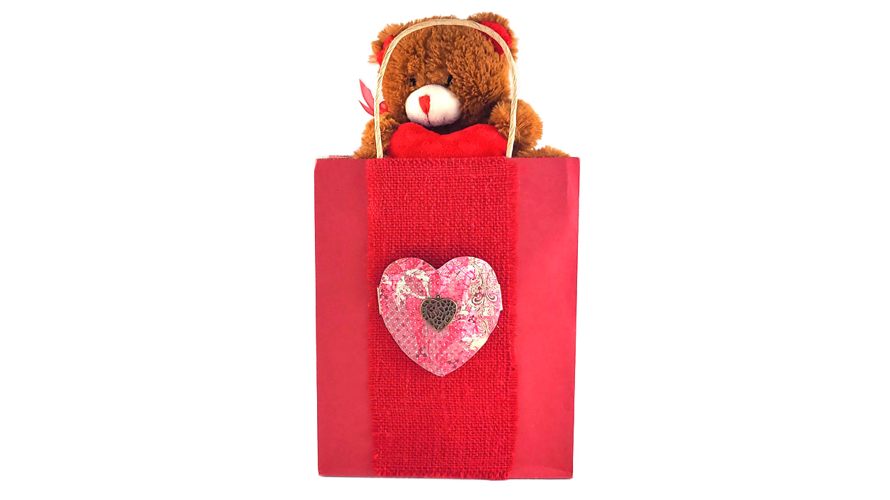 Red Valentine's Day gift bag decorated with a red burlap strip and a pink paper heart ornament, featuring a brown teddy bear plushy peeking out of the top as a tissue paper alternative.