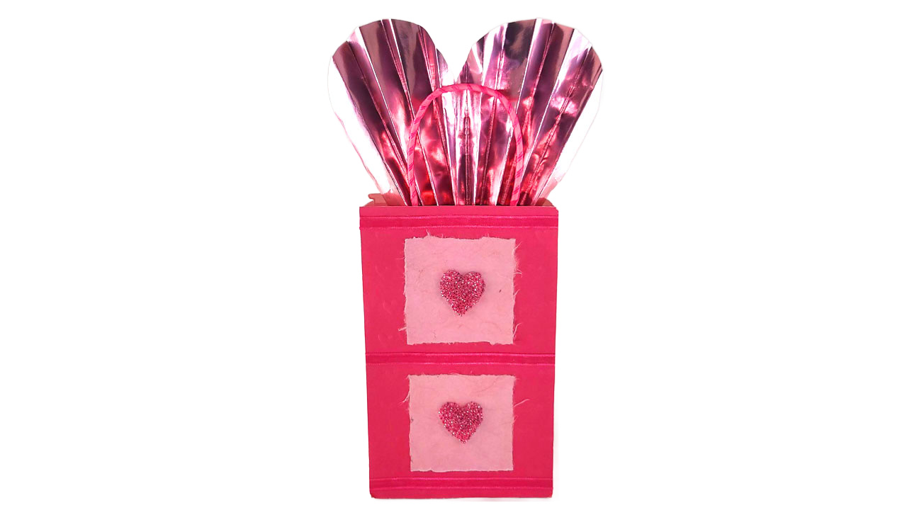 Pink gift bag with fabric hearts featuring a metallic pink wrapping paper fan shaped like a heart sticking out the top.