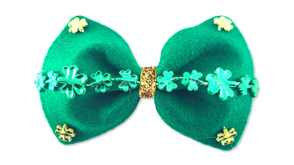 A high-end DIY St. Patrick’s Day bow tie made from green felt with a gold glitter ribbon center. It is decorated with a row of green sequin shamrocks and gold shamrock accents in the corners, showcasing an original design.