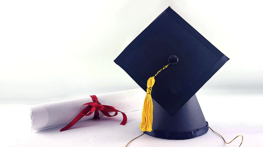 Graduation cap with yellow tassel next to a rolled diploma tied with a red ribbon.