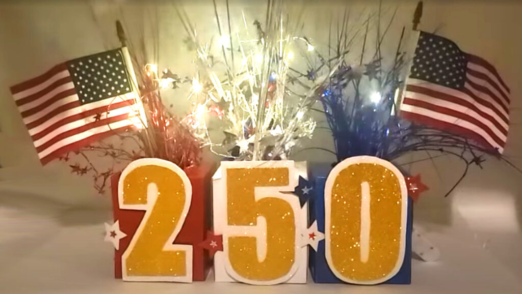 Patriotic July 4th centerpiece featuring glittery “250” numbers, American flags, and red, white, and blue streamers arranged on a festive display.