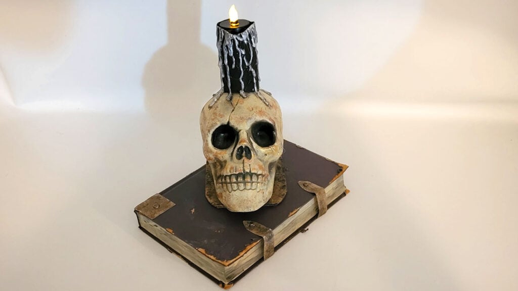 Black faux wax‑dripped Halloween candle sitting on a skull and aged book, made using a paper towel roll for spooky DIY decor.