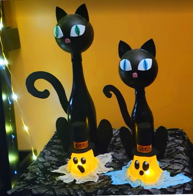 Two handmade black cat Halloween decorations standing on glowing ghost bases with festive lights in the background.