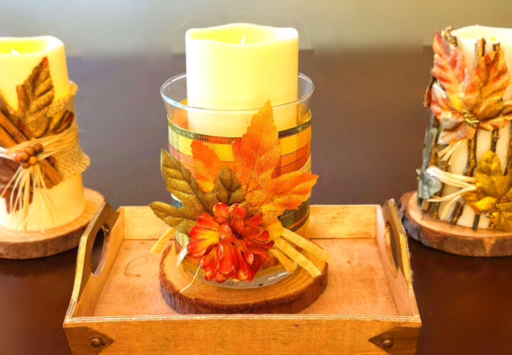 Three rustic DIY fall candles sit on a wooden tray. One is wrapped in cinnamon sticks, another has a plaid ribbon and autumn leaf, and the third is decorated with twigs and gold leaves, creating a cozy autumn scene.