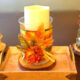 Three rustic DIY fall candles sit on a wooden tray. One is wrapped in cinnamon sticks, another has a plaid ribbon and autumn leaf, and the third is decorated with twigs and gold leaves, creating a cozy autumn scene.