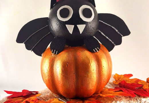 This Adorable DIY Dollar Tree foam pumpkin bat on a small hay bale. The cute black bat sits on a metallic orange pumpkin, surrounded by autumn leaves, creating a perfect Halloween decor scene.