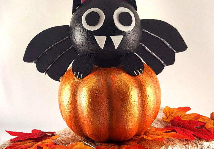 This Adorable DIY Dollar Tree foam pumpkin bat on a small hay bale. The cute black bat sits on a metallic orange pumpkin, surrounded by autumn leaves, creating a perfect Halloween decor scene.