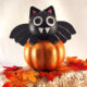 This Adorable DIY Dollar Tree foam pumpkin bat on a small hay bale. The cute black bat sits on a metallic orange pumpkin, surrounded by autumn leaves, creating a perfect Halloween decor scene.