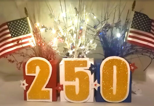 Patriotic July 4th centerpiece featuring glittery “250” numbers, American flags, and red, white, and blue streamers arranged on a festive display.