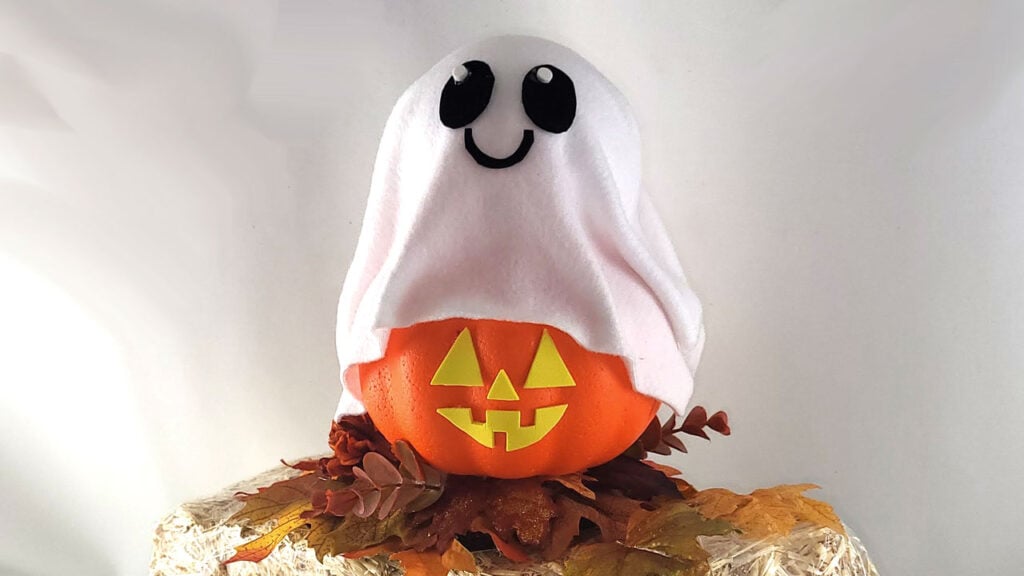 A cute no‑carve ghost pumpkin made from a Dollar Tree foam pumpkin covered with white fabric, decorated with black ghost eyes and a smiling mouth, sitting on fall leaves.
