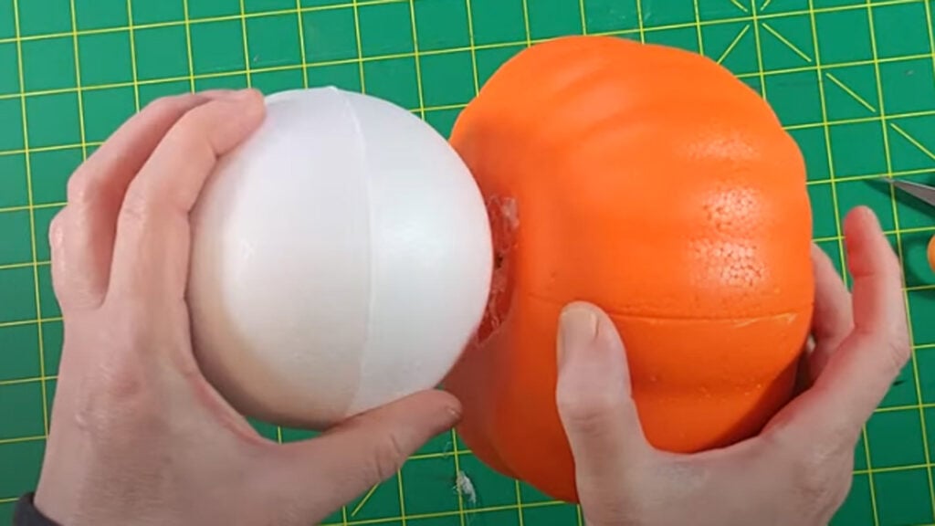 Hands holding a white Styrofoam ball and an orange foam pumpkin on a green cutting mat, showing the first step of making a Dollar Tree ghost pumpkin craft.