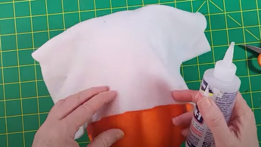 Hands draping white fabric over a foam pumpkin and Styrofoam ball on a green cutting mat while adding glue to create the ghost shape for a Dollar Tree ghost pumpkin craft.