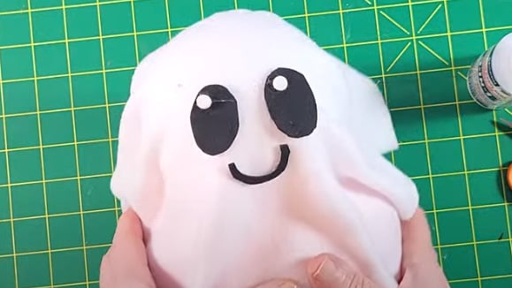 Hands holding a small ghost craft made from white fabric with black foam eyes and a smiling mouth on a green cutting mat.