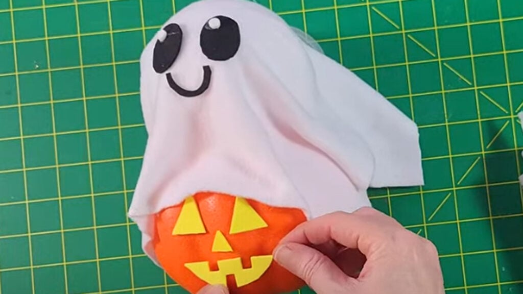 Hand holding a small pumpkin craft with a yellow jack‑o‑lantern face and a white ghost cloth with black eyes and a smile on a green cutting mat.