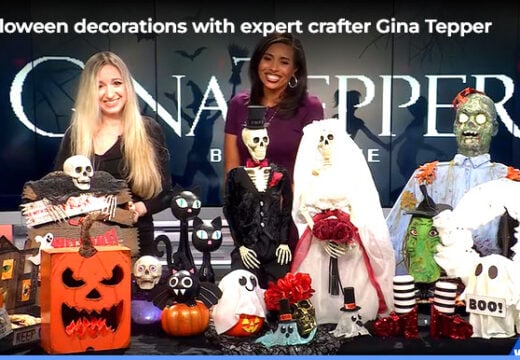 Gina Tepper and a WBAL-TV host standing behind a display of DIY Halloween decorations, including a flaming pumpkin, a haunted house, skeletons, a ghost, and other spooky crafts.
