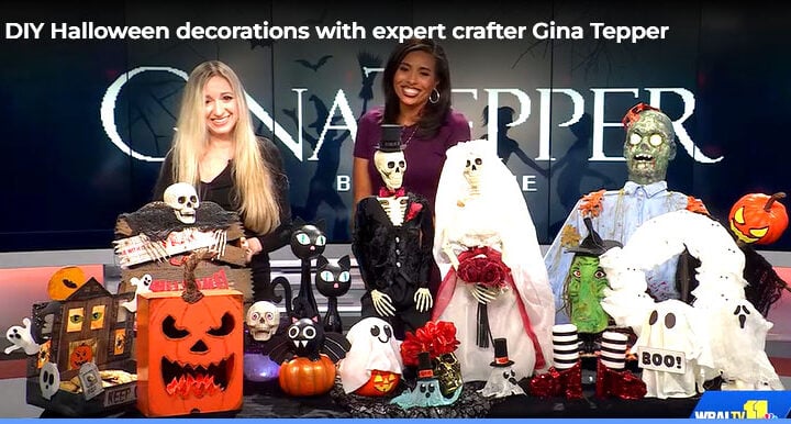 Gina Tepper and a WBAL-TV host standing behind a display of DIY Halloween decorations, including a flaming pumpkin, a haunted house, skeletons, a ghost, and other spooky crafts.