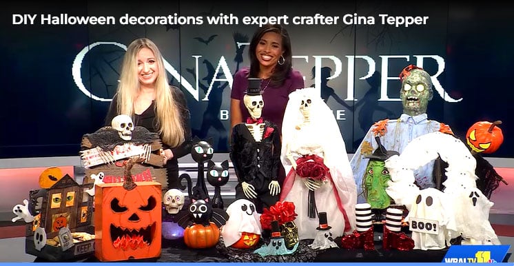 Gina Tepper and a WBAL-TV host standing behind a display of DIY Halloween decorations, including a flaming pumpkin, a haunted house, skeletons, a ghost, and other spooky crafts.
