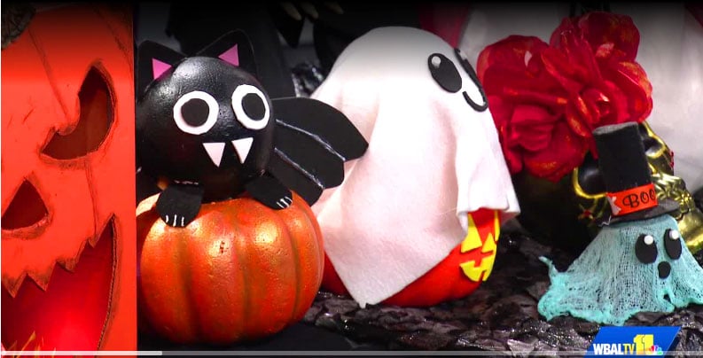 A display of handmade Halloween decorations featuring an orange jack-o'-lantern, a black cat sitting on a pumpkin, a white ghost, a red decorative flower, and a small blue ghost.